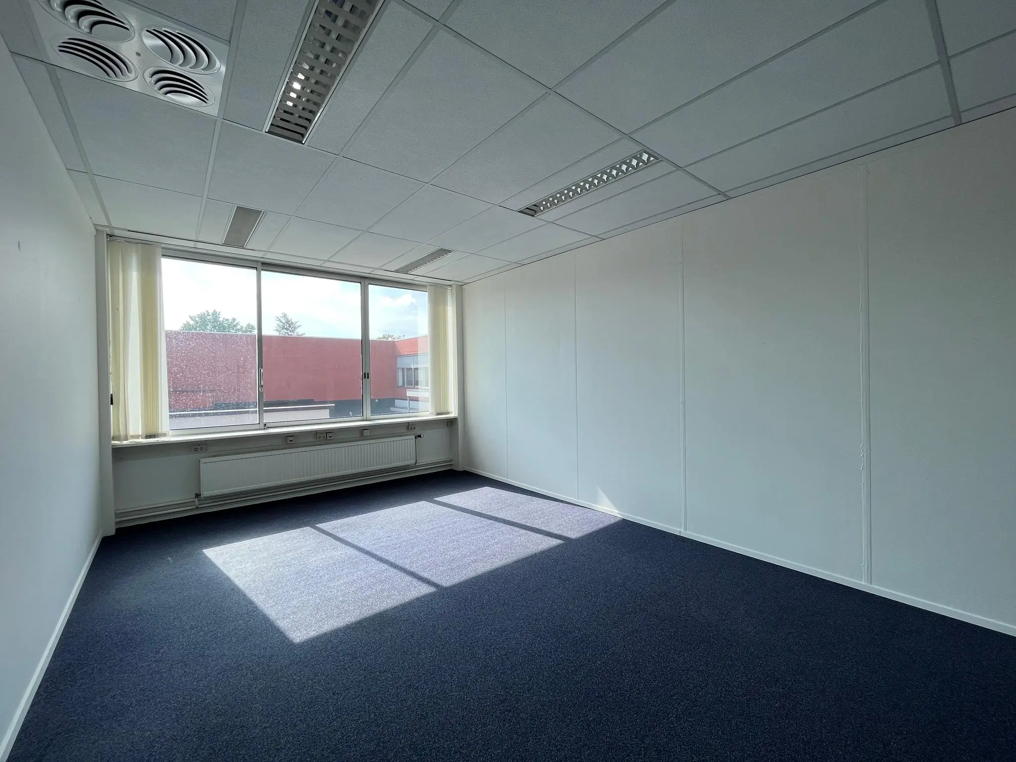 Leeg, licht kantoor met blauw tapijt aan de Jan van Eycklaan, met groot raam en systeemplafond.